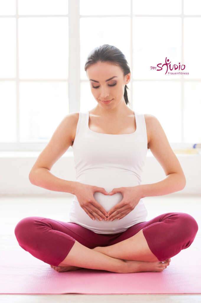 eine junge, schwangere Frau sitzt im Lotussitz auf ihrer YogamatteHänden vor ihren Bauch.